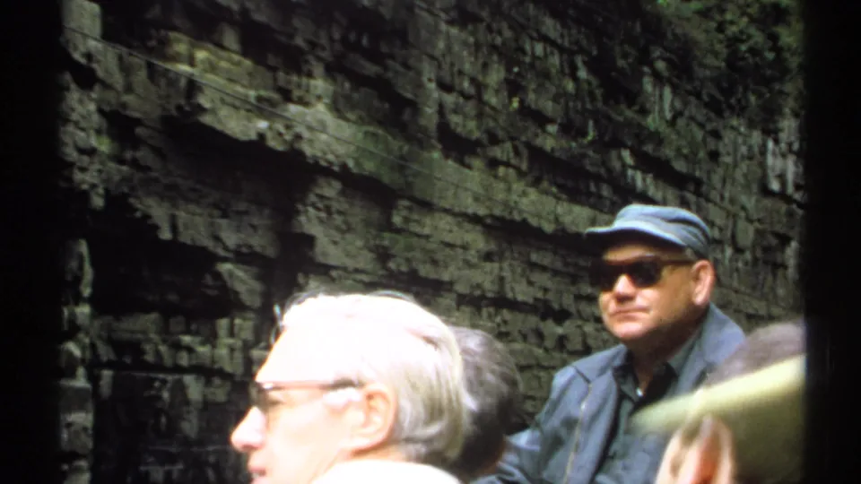 1971 New York 8mm frame showing adult tourists paused inside a layered rock gorge, including a man in a blue cap and sunglasses.