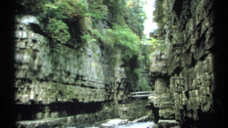 The 1971 New York home-movie view looks down a narrow gorge with rock walls, greenery above, railings, and a stream below.