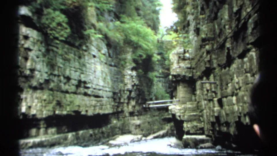 A handheld 8mm view of the New York gorge shows water below, rock ledges, greenery, railings, and a partial tourist at frame edge.
