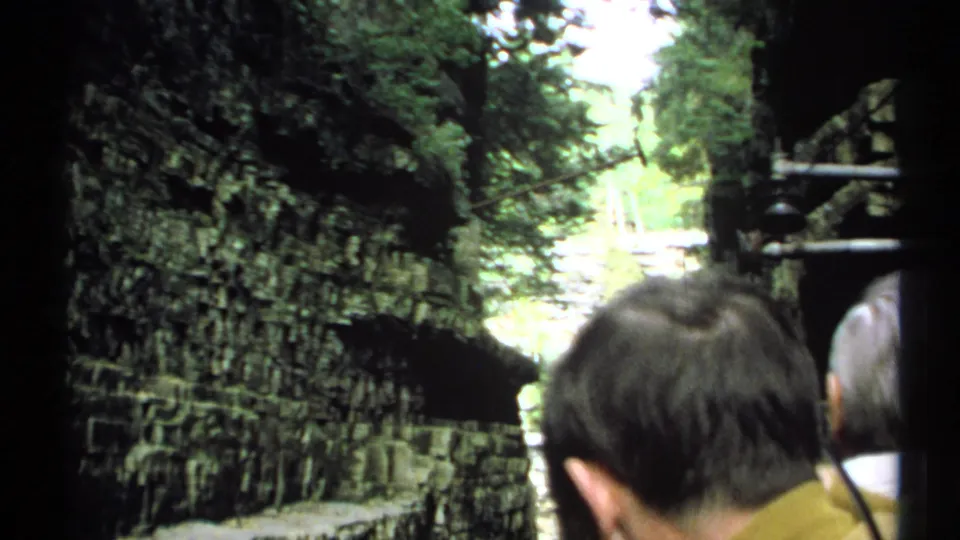 1971 New York home-movie frame showing visitors in the foreground beside dark limestone walls and a sunlit stream corridor.