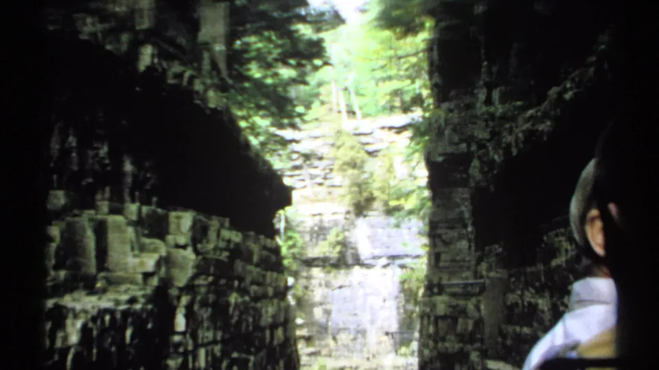 1971 New York 8mm frame looking through shadow toward a rocky river canyon with layered stone walls, water, and green foliage.