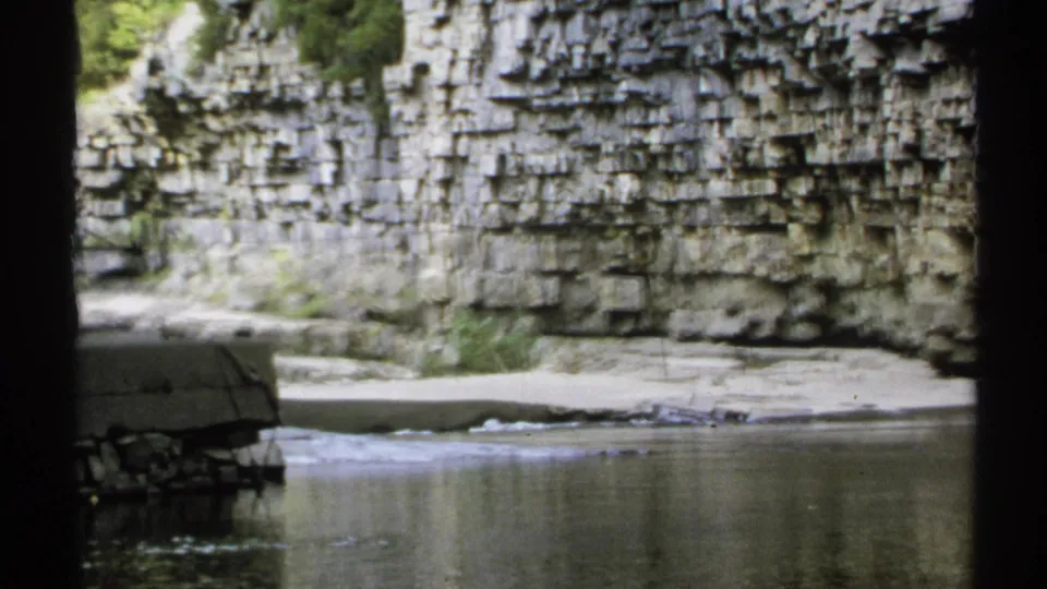 1971 New York 8mm view across still river water toward a layered canyon wall and low stone or concrete edge.