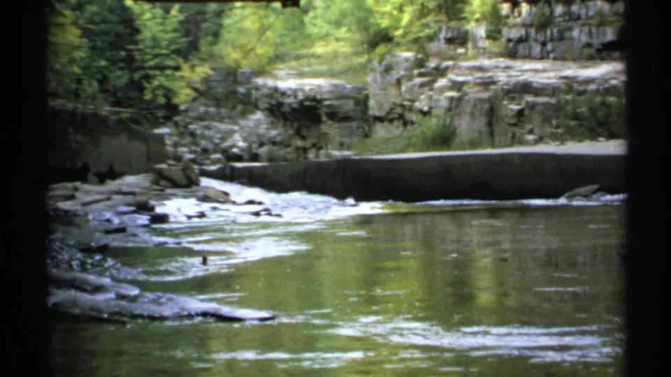1971 New York 8mm frame showing a quiet river bend, layered rock ledges, shallow water, and dense green foliage overhead.