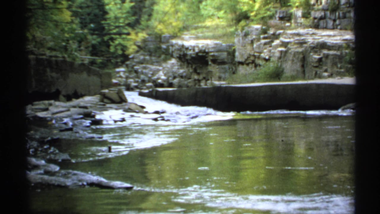 Rocky river with a low spillway and forested cliffs in the background.