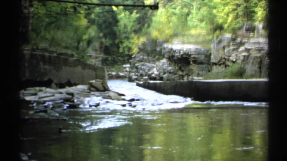 1971 New York 8mm view across a river toward a small cascade, stone ledges, and dense green trees.