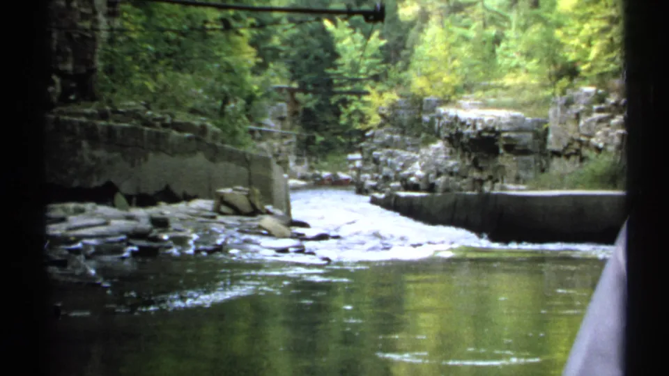 1971 New York 8mm frame of a calm river foreground with white water over rocks and wooded cliffs beyond.