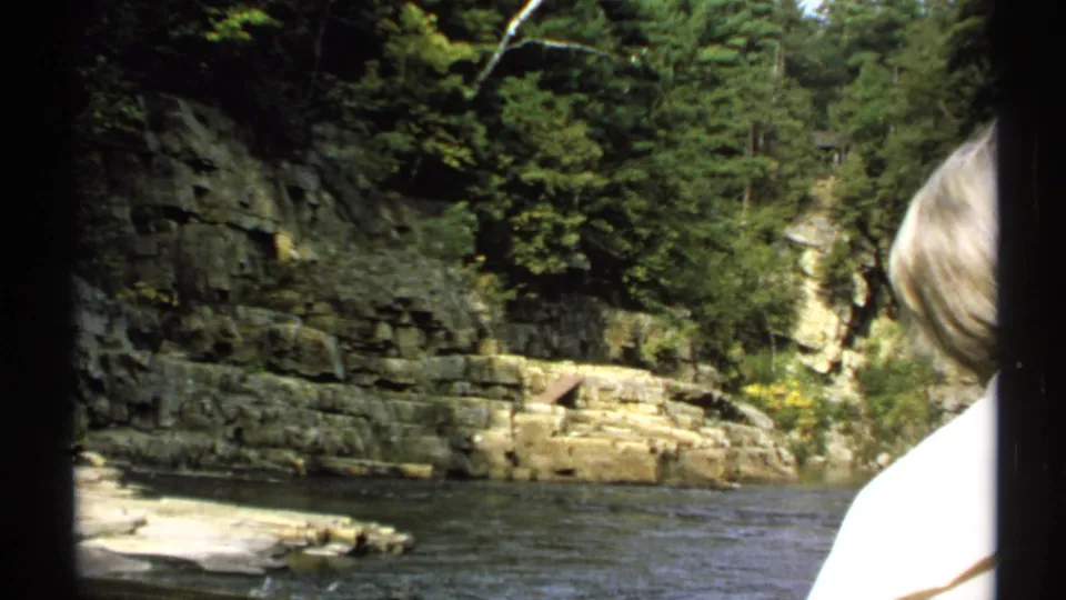 1971 New York 8mm frame with a light-haired observer seen from behind at the river, facing wooded cliffs and stone ledges.