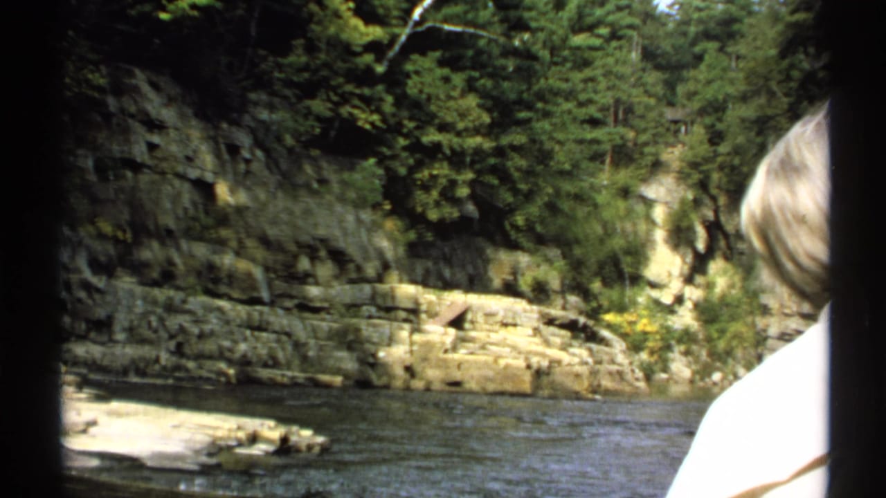 River passing layered rock banks and trees, with a light-haired person at the right edge.