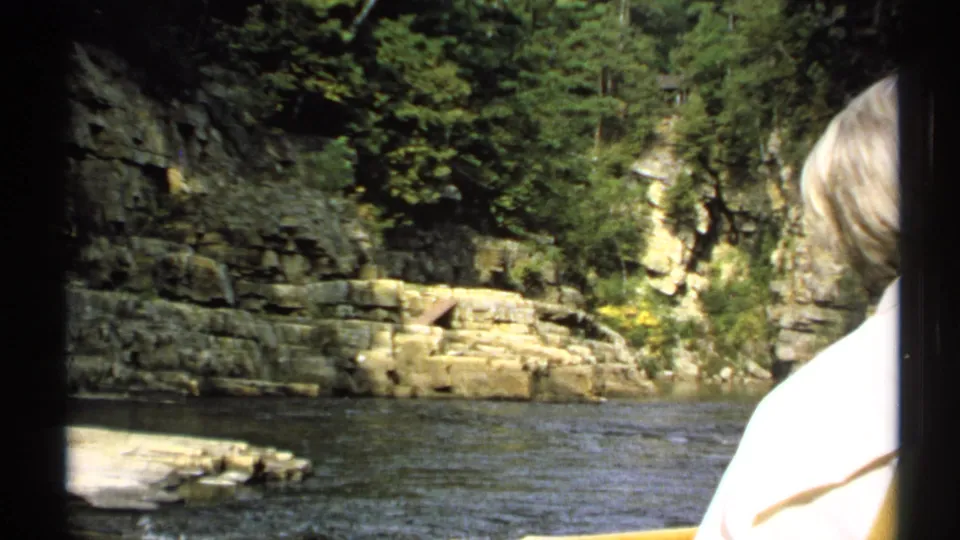 1971 New York home-movie frame showing a wooded rock bank across the river, framed by dark film borders and a foreground passenger.
