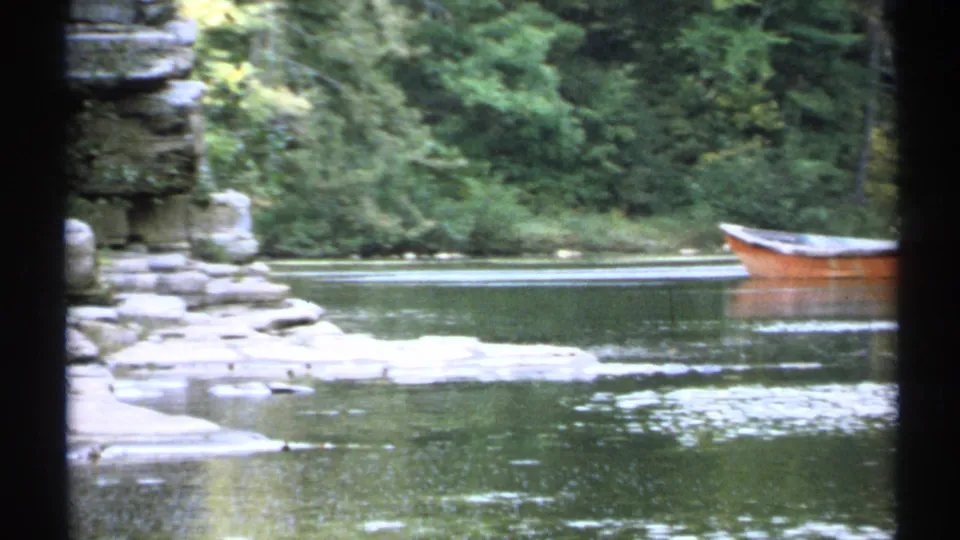 1971 New York 8mm frame of a calm lake, stone shoreline, forested bank, and orange rowboat near the edge of the water.