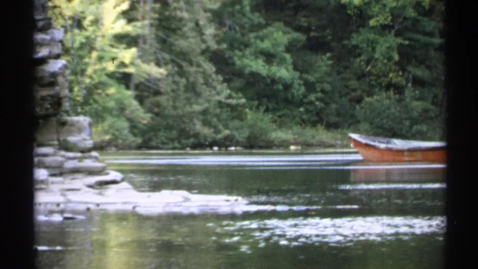 1971 New York home-movie frame showing an orange rowboat on still water, with a rocky bank and dense green trees behind it.