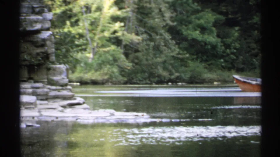 1971 New York 8mm frame with a partial orange rowboat at the edge, rippling lake water, rocks, and a forested shoreline.
