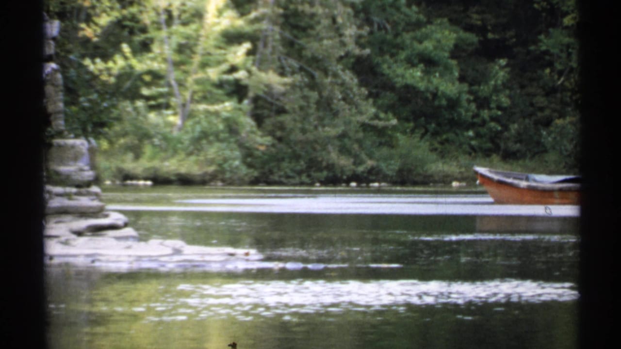 Brown rowboat on a calm pond beside trees and a stone wall, partly blocked by dark foreground shapes.