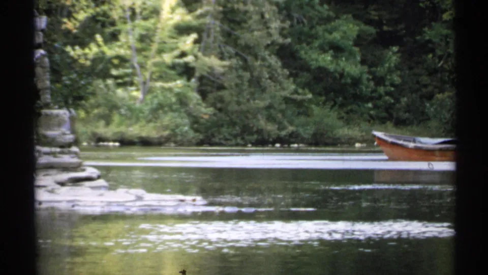 1971 New York 8mm frame showing a brown rowboat on still water near trees and a stone retaining wall, viewed through a partial obstruction.