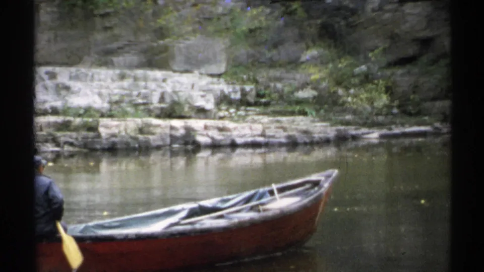 1971 New York home-movie frame with a grey-jacketed rower beside a brown rowboat on calm pond water near a wooded stone bank.