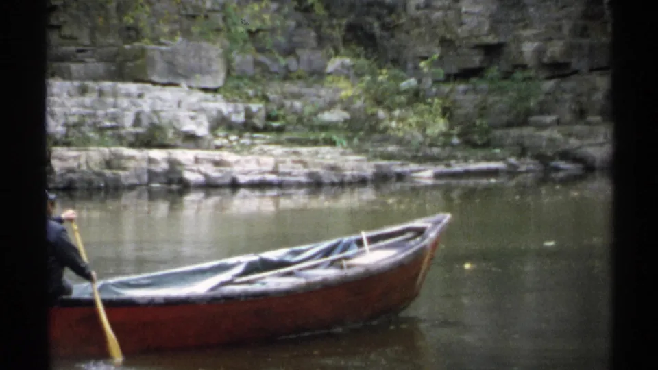 A 1971 New York 8mm frame shows the rower partly cropped at left, using a yellow oar beside a brown rowboat on still water.