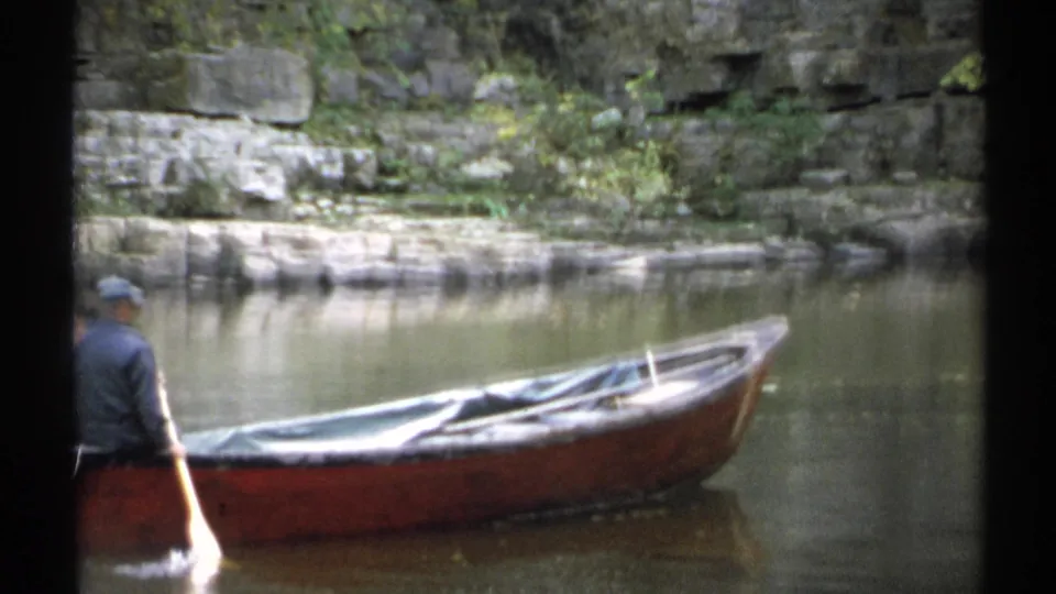 1971 New York frame showing a grey-jacketed rower with a yellow oar next to a brown rowboat, with still water and stonework behind.