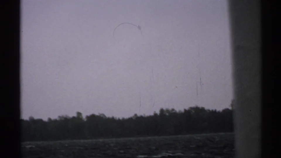 1971 New York 8mm landscape with a distant tree line, rocky shoreline, water, and dark window framing.