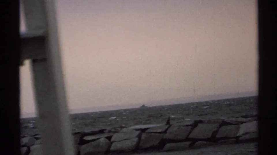 The 1971 coastal view looks past a pale structural post toward dark rocks, choppy water, and a small boat near the horizon.