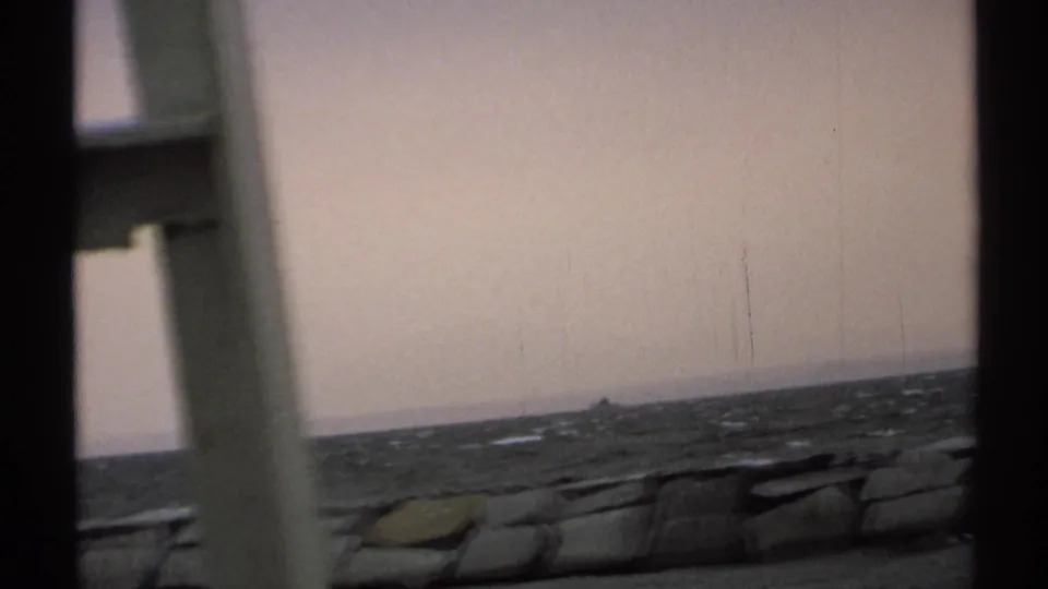 A subdued 8mm frame shows the New York coast with choppy water, a low stone shore edge, and a small boat under a pale sky.