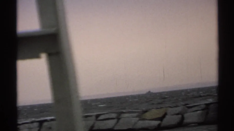 1971 8mm New York boat view framed by pale structure, with open water, low shoreline, and a distant dark maritime marker.