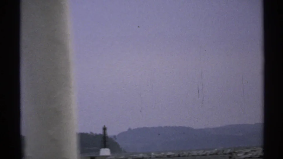 1971 New York home-movie frame showing a hazy shoreline and dark vertical marker beyond pale boat framing.