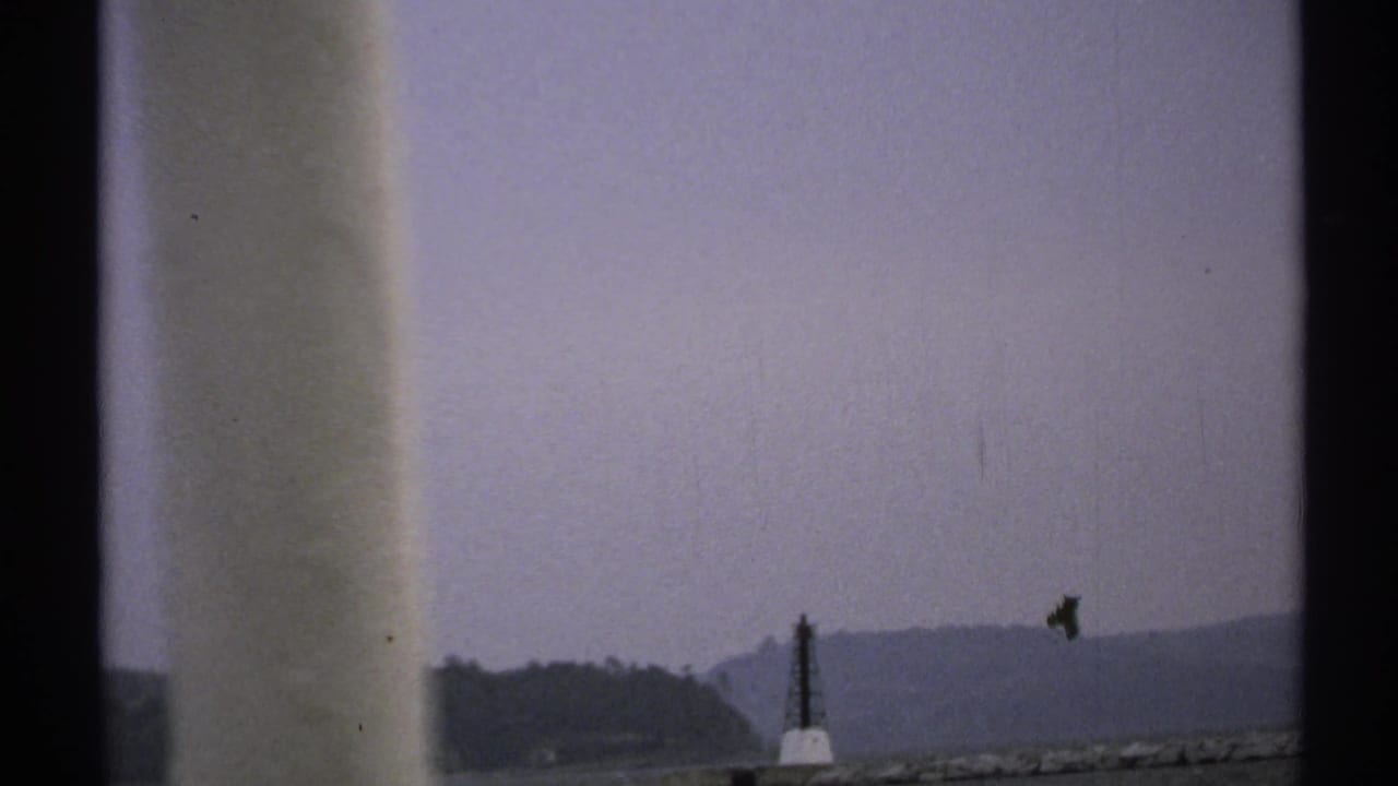 1971 New York home-movie frame showing water, distant shoreline, and a small boat, partly obscured by dark film-frame borders.