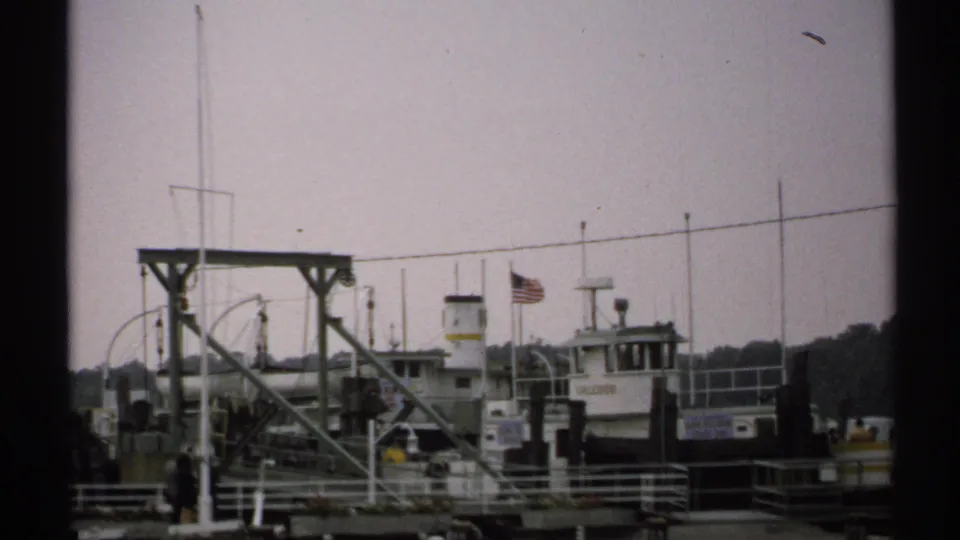 1971 New York waterfront frame with a docked ferry, American flag, metal loading structure, pier railings, and distant shoreline.