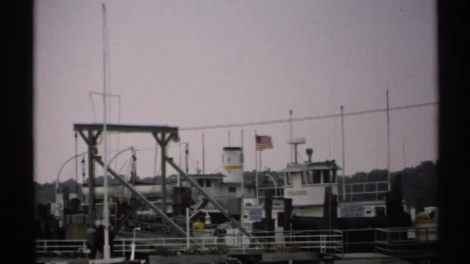 1971 New York home-movie frame showing a ferry at a waterfront pier, metal loading framework, railings, and a distant person standing near the dock.