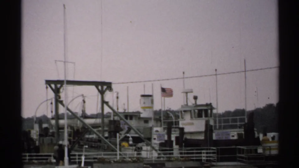 1971 New York frame showing the docked ferry, American flag, metal pier structure, railings, and soft overcast waterfront light.