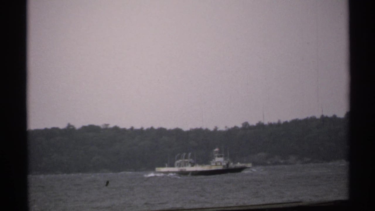 1971 home-movie frame of a distant boat on a river or broad waterway, seen through dark window framing with trees in the background.