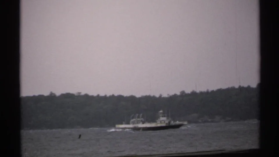 1971 home-movie frame of a distant boat on a river or broad waterway, seen through dark window framing with trees in the background.