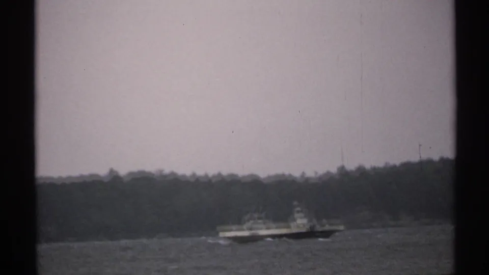 Distant boat on a broad waterway in 1971 footage, with a tree-lined shore and dark window or film-frame borders visible.