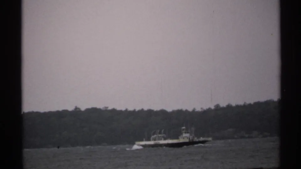 Home-movie view of a distant vessel crossing choppy water, with wooded shoreline and heavy dark frame borders visible.