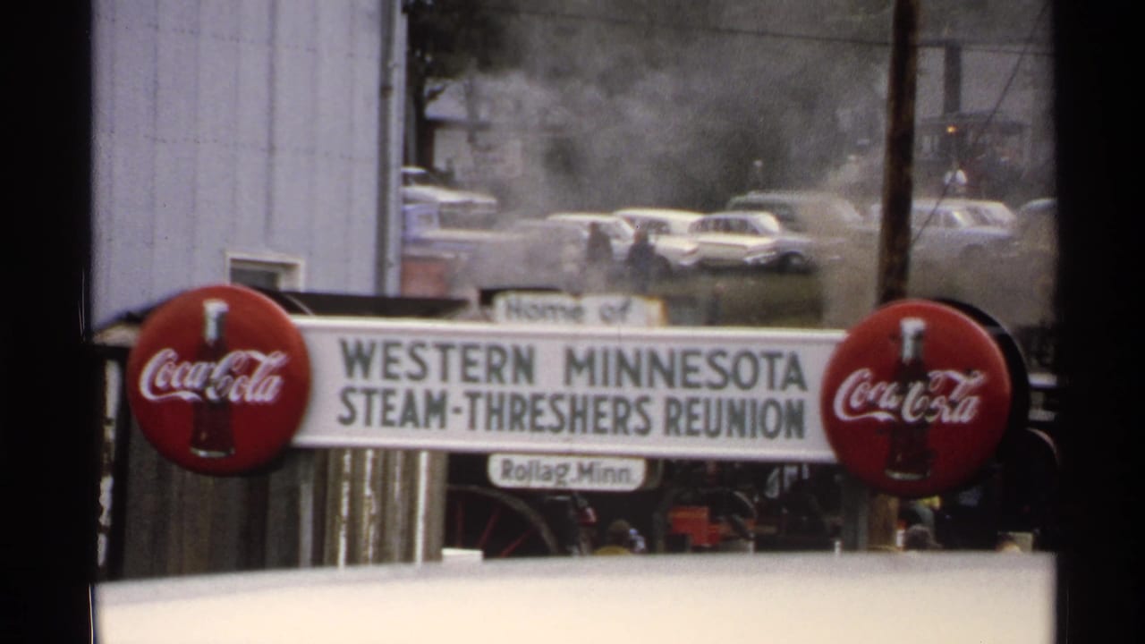 Western Minnesota Steam-Threshers Reunion sign flanked by red Coca-Cola signs with cars and smoke behind it.
