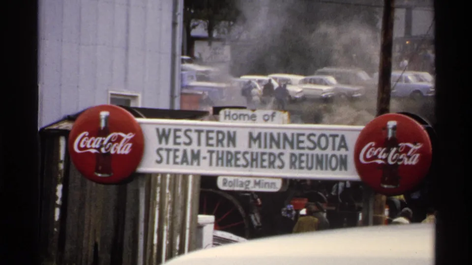 The Rollag reunion sign remains centered, with Coca-Cola signs on both sides and faint visitors and parked cars visible beyond drifting steam.