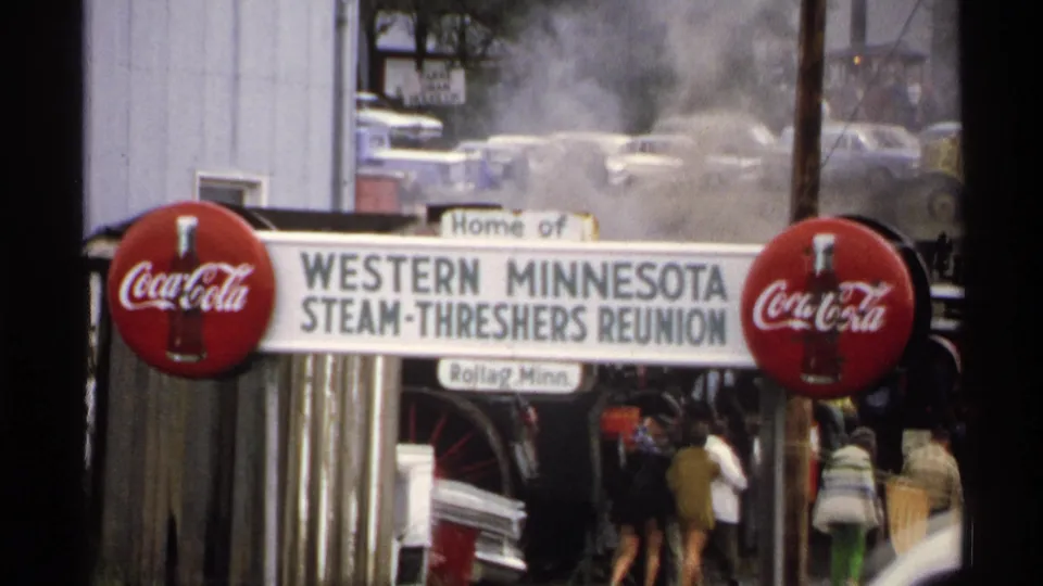 Visitors in 1970s clothing move near the Western Minnesota Steam-Threshers Reunion sign, with red Coca-Cola signs and steam haze behind them.