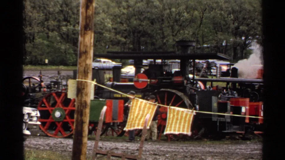 A large steam engine stands at the Rollag reunion grounds, with red wheels, a striped barrier cloth, trees, parked cars, and steam smoke nearby.