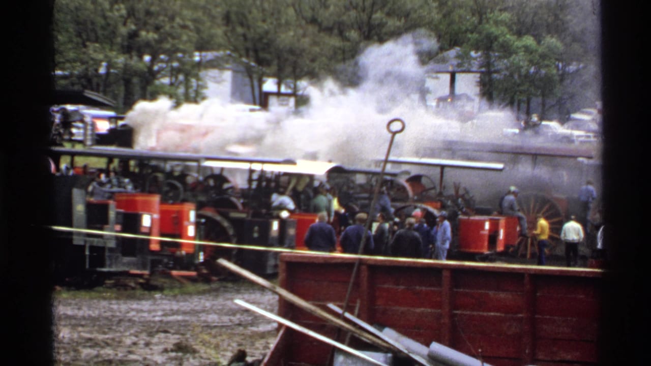 Steam tractors with smoke surrounded by spectators in an outdoor field.