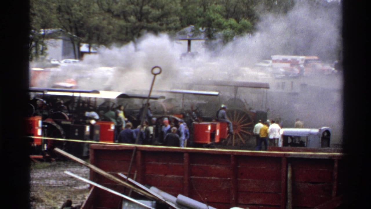 1971 Rollag, Minnesota 8mm frame showing steam-powered tractors smoking while spectators stand near red machinery at an antique tractor show.
