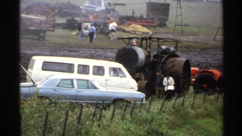 1971 Rollag, Minnesota show grounds with a white van, light blue sedan, rusted steam engine, and visitors walking near farm equipment.