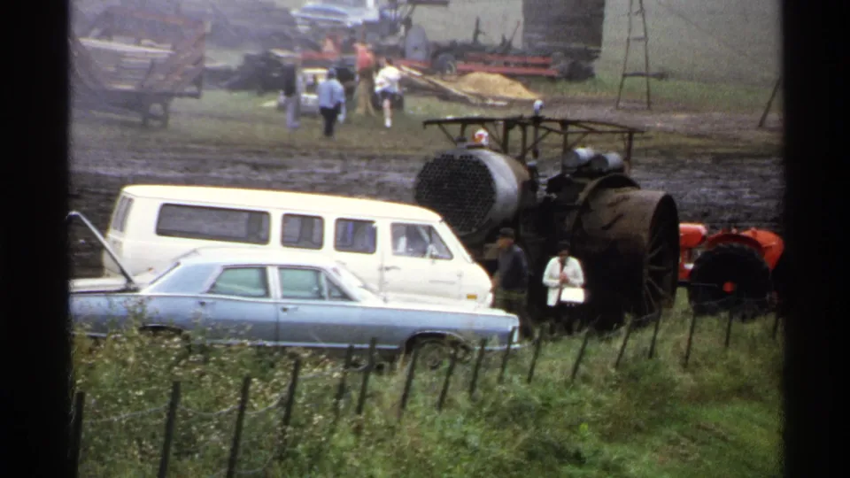 1971 Rollag, Minnesota frame with parked period vehicles, a dark steam engine, fence posts, and visitors at the antique tractor show.