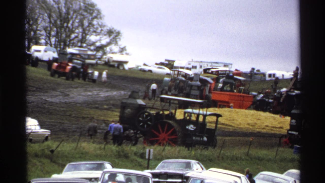 Classic cars in the foreground with antique tractors and visitors on a grassy hillside.