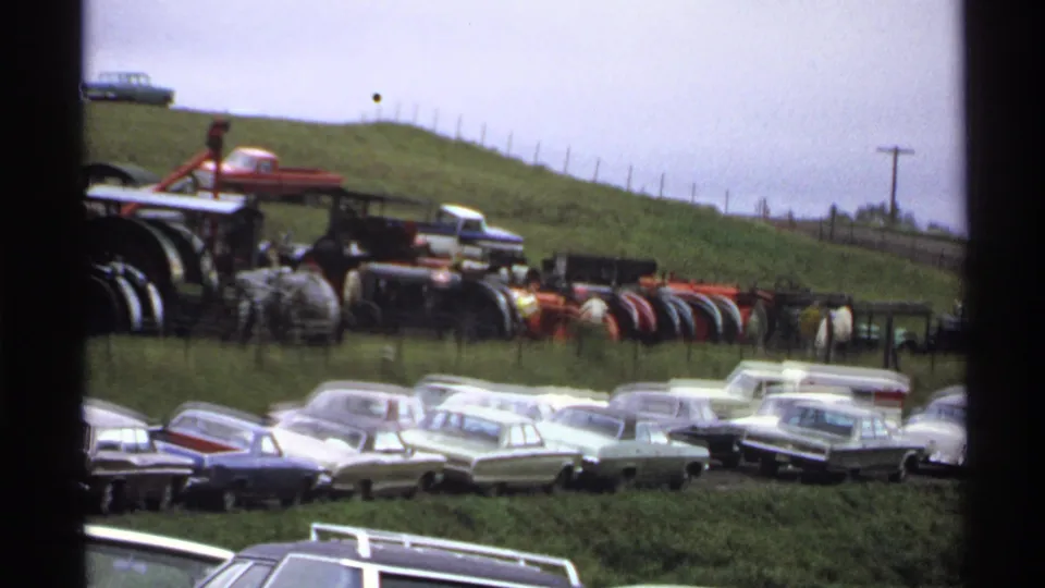 1971 Rollag, Minnesota fair view with parked American cars below a line of vintage tractors and machinery on the slope.