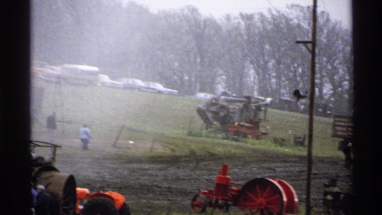 Steam tractor display in a muddy field with parked cars on a grassy hill.