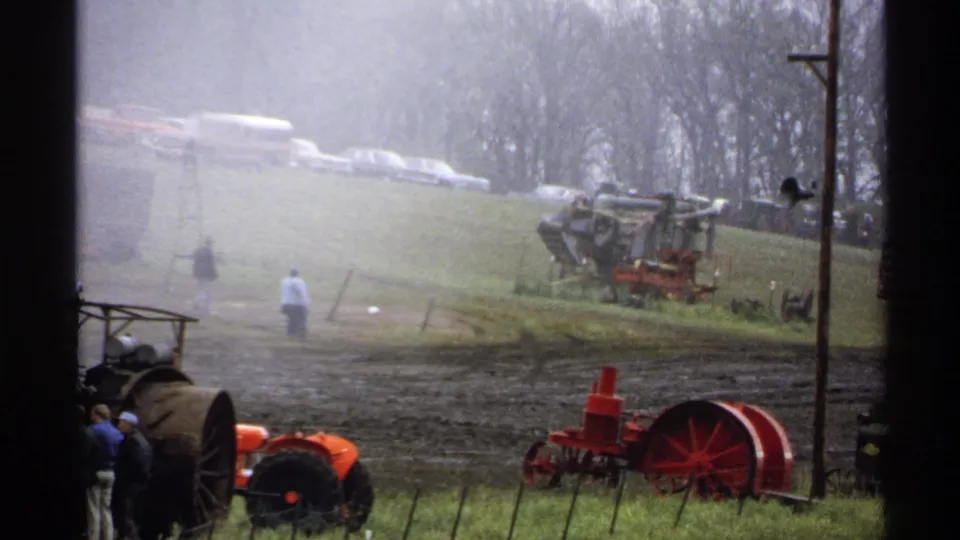At the Rollag, Minnesota fairground, visitors stand near vintage farm machinery while classic cars line the grassy hill behind them.
