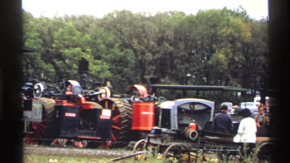 1971 Rollag fair footage frame with visitors moving near red and black steam machinery against a dense tree backdrop.