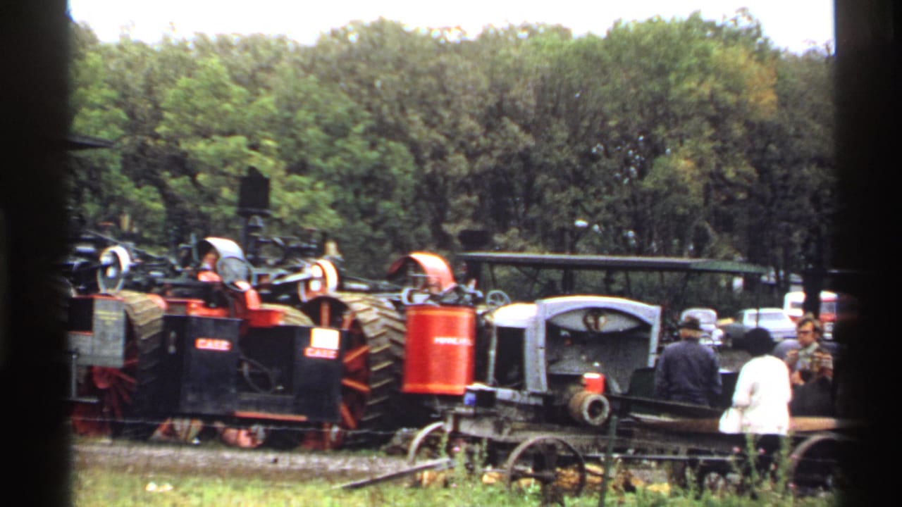 1971 Rollag, Minnesota home-movie frame showing steam tractors on display with visitors standing near a wagon and trees behind them.