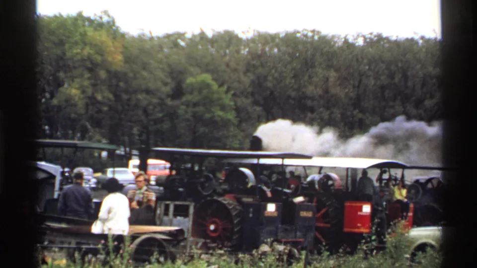 1971 Rollag, Minnesota home-movie frame showing parked steam tractors, a few spectators, and white exhaust against a wooded background.