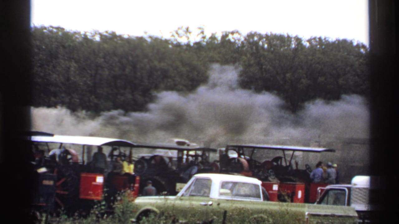 Steam traction engines with spectators, smoke, and a green and white pickup in the foreground.